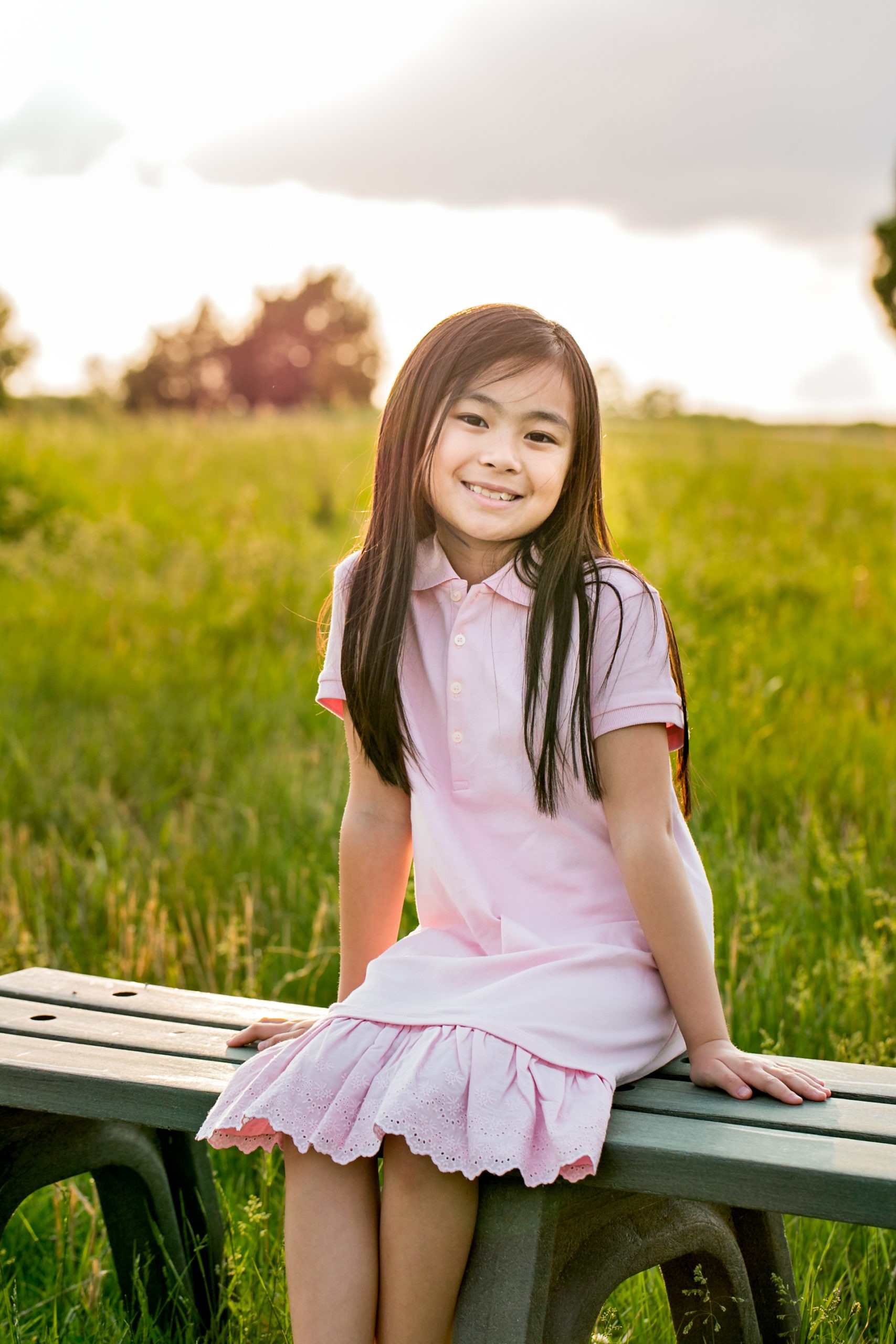 Smiling girl in a pink dress sitting on a bench in a grassy field, representing the friendly and welcoming environment of Moon Signature Dentistry for children's dental care in Olathe.