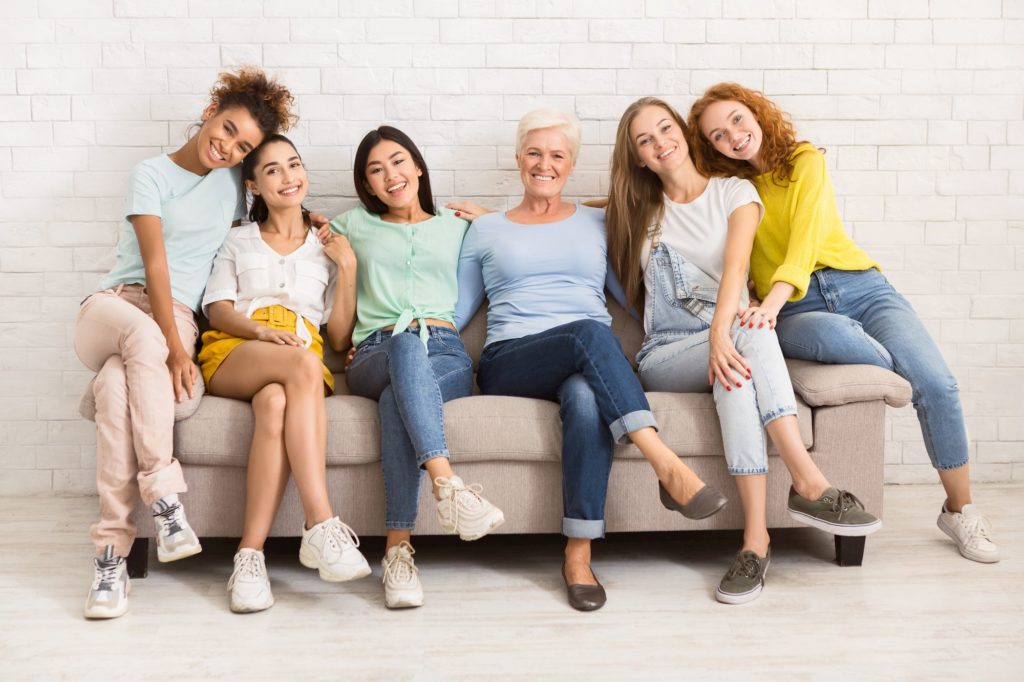 Group of six women of diverse ages and styles smiling together on a couch, representing a welcoming atmosphere for family and general dentistry services at Moon Signature Dentistry in Lenexa.