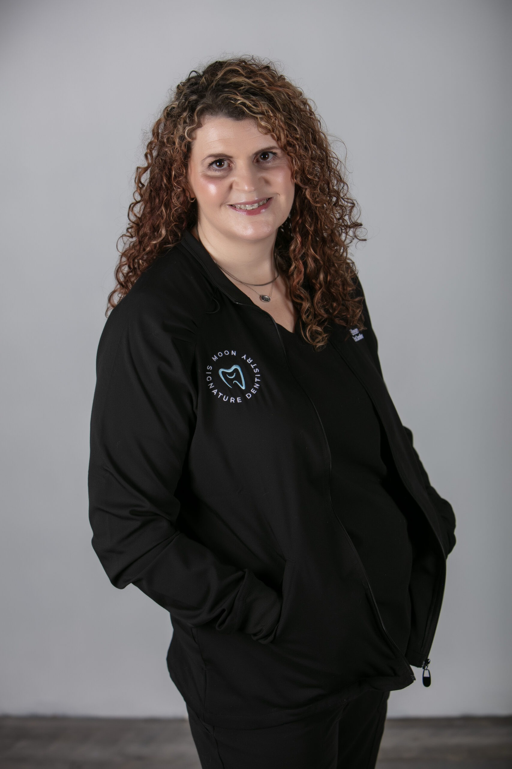 Shanan, Clinical Coordinator at Moon Signature Dentistry, smiling in a black dental jacket, showcasing the practice's logo, in a neutral background.